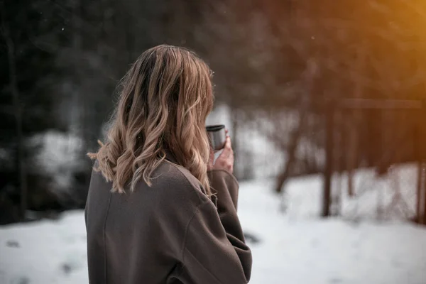 Blonde girl drinks hot beverage from thermos cup in winter dramatic forest. Getting warm during snowfall