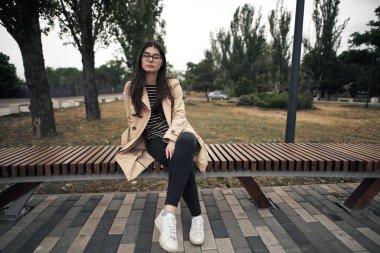 Genç esmer kız bankta oturur ve kameraya bakar. Kahverengi trençkotlu modern iş kadını. Gözlük takan kadınlar