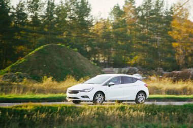 Modern beyaz kompakt hibrit araba sonbahar ağaçlarının önündeki asfalt yolda gidiyor.