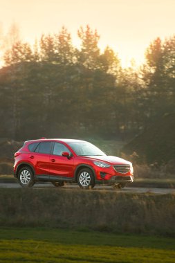 Parlak sonbahar yeşillikleri ve ormanlarla çevrili asfalt bir köy yolunda hızla ilerleyen bir SUV.