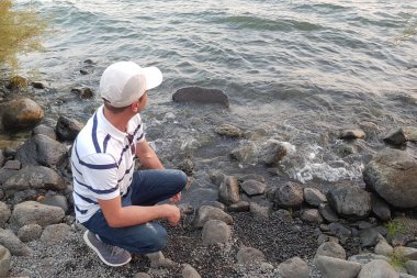 Beyaz beysbol şapkalı, beyaz mavi çizgili tişörtlü ve mavi kotlu bir adam gölün kayalık sahilinde oturur ve suyun içine bakar.