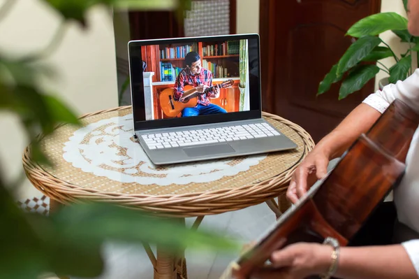 Woman learning to play guitar with remote class or tutorial using a computer in a lovely room of the house full of plants. Technology and learning from home.