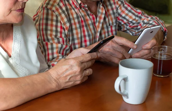 Elderly couple using modern mobile phones at home while enjoying cups of hot drinks. Concept of elderly people and technology, personal development...