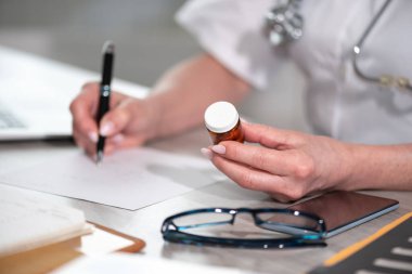 Kadın doktor ilaç şişesine bakıyor ve reçete yazıyor.