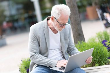 Olgun bir iş adamı dışarıda bir bankta oturuyor ve dizüstü bilgisayar kullanıyor. 