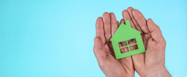 Hands holding a green house light on light blue background; Concept of eco friendly house