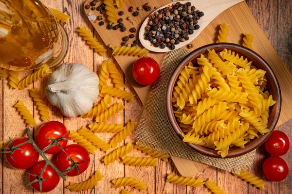 On a wooden table, pasta in a ceramic bowl, cherry tomatoes, garlic, olive oil, a mixture of peppers in a wooden spoon.