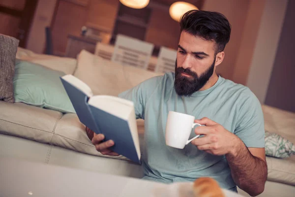 Jovem Está Sentado Sofá Lendo Livro Enquanto Segura Xícara Café — Fotografia de Stock