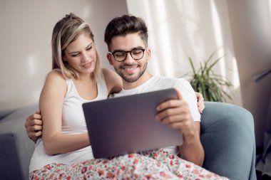 Dizüstü bilgisayar kullanarak ve evde kanepede otururken gülümseyen güzel genç çift.