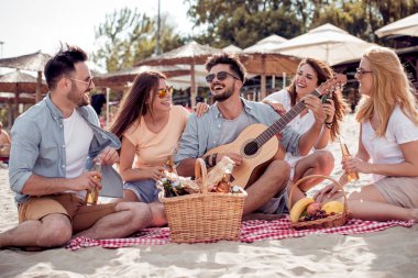 Sahilde kum üzerinde oturan arkadaşlar. Genç adam gitar çalıyor.