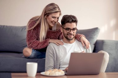 Güzel genç çift evde kanepede otururken gülümseyen, dizüstü bilgisayar kullanarak.