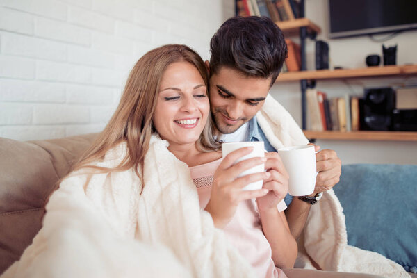 Cute couple relaxing on couch under blanket at home in the living room and drink coffee.