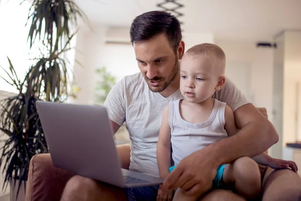 Family, fatherhood, technology and people concept - happy father and little son with laptop sitting on armchair at home.