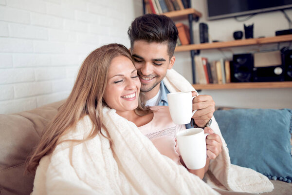 Cute couple relaxing on couch under blanket at home in the living room and drink coffee.