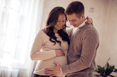 Mutlu hamile kadın kocası ile tutarak ve göbek bebek dinleme ve evde oturma odasında duran.