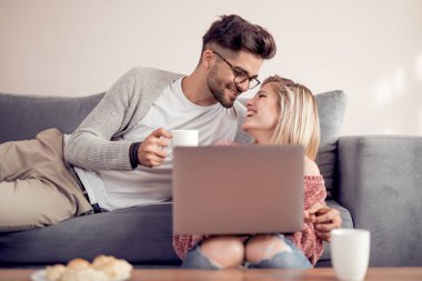 Çift kanepede oturan, kahve içme ve dizüstü evde kullanma.
