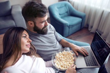 Mutlu genç çift evde laptop film izlerken patlamış mısır ile kanepede oturan.