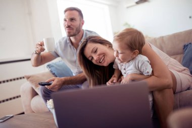 Evde bir dizüstü bilgisayar kullanarak neşeli bir aile portresi. Baba elinde bir uzaktan kumanda ve Tv izlerken.