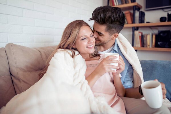 Cute couple relaxing on couch under blanket at home in the living room and drink coffee.