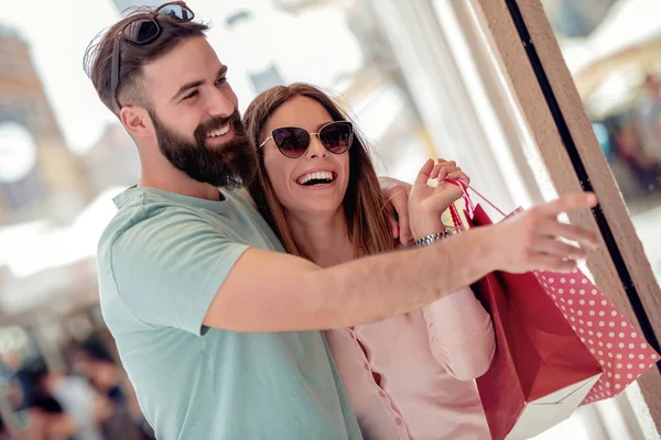 年轻的情侣带着购物袋看着商店 男人正在橱窗上展示他的女朋友 — 图库照片