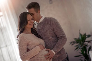 Yakışıklı bir adam ve güzel hamile karısı evlerinin penceresinin yanında durup gülümsüyorlar..