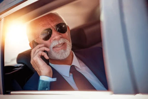 mature senior businessman in suit and glasses with mobile phone riding on back seat of car