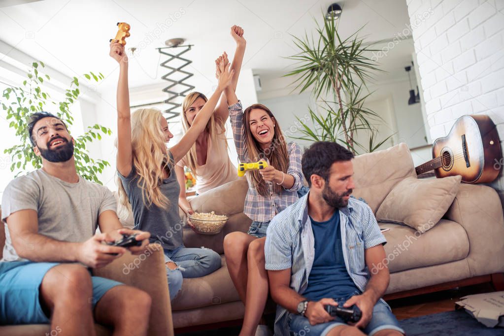 Excited friends playing video games at home, sitting on sofa and enjoying together.