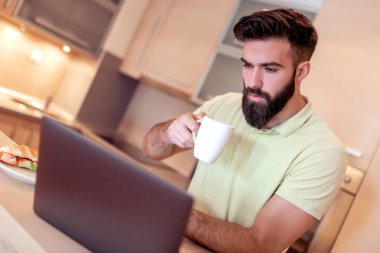 mutlu bir adam mutfakta kahve yudumlarken onun laptop üzerinde çalışma.