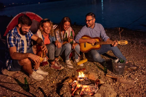 Group of friends sitting by the fire at night, grilling sausages and having great time on the beach.