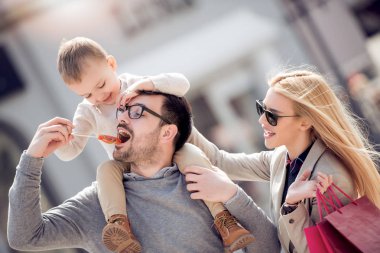 Baba oğlu Anne yanında güler onun omuzlarında taşır. Güneşli günde şehirde oturan aile.