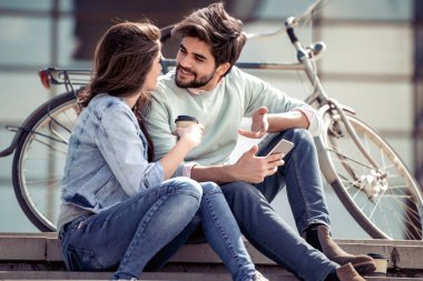 Genç, gülümseyen bir çift merdivenlerde otururken cep telefonu kullanıyor ve kahve içiyor..