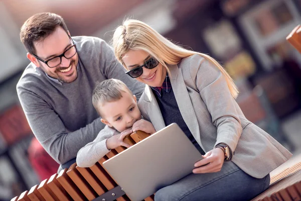 Happy family and digital technology. Young parents and little kid using laptop while sitting in the city park.