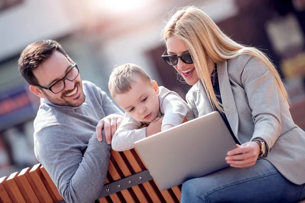 Happy family and digital technology. Young parents and little kid using laptop while sitting in city park.