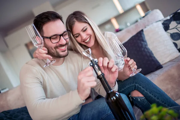Oturma odasında kanepede oturan genç romantik çift şarap içmek ve aşık tadını çıkarın. Mutlu bir adam bir şişe şarap açıp.