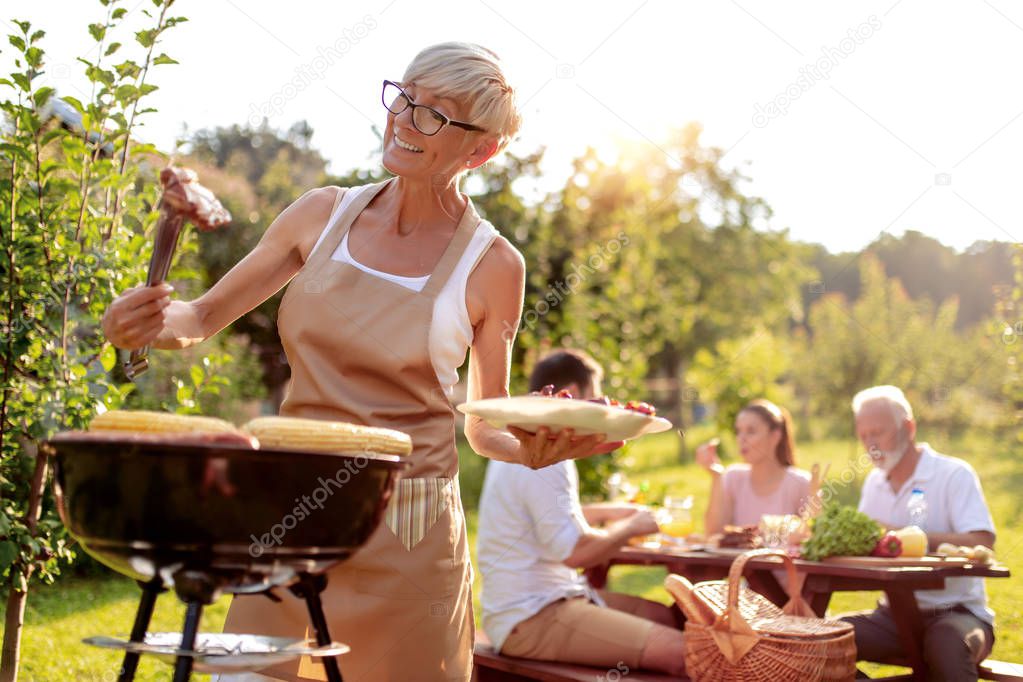 Food, people and family time concept-senior woman cooking meat on barbecue grill at summer garden  party.