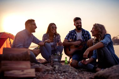 Genç çekici arkadaşlar grubu nehir yanında oturuyor, gitar çalıyor ve eğleniyor.
