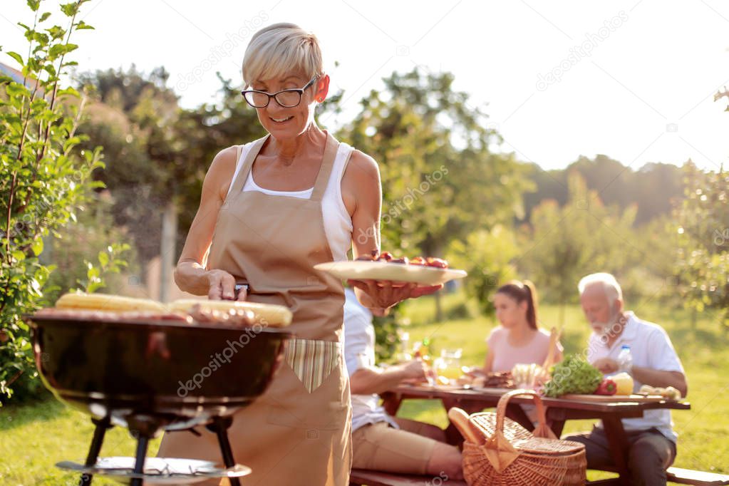 Food, people and family time concept-senior woman cooking meat on barbecue grill for her family.