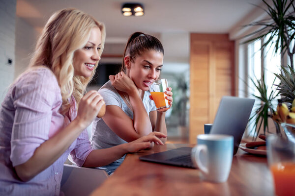 Две красивые женщины, сидящие за столом на кухне и пользующиеся ноутбуком
.