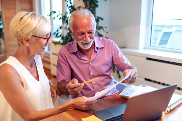 Senior couple at home using laptop and holding document.