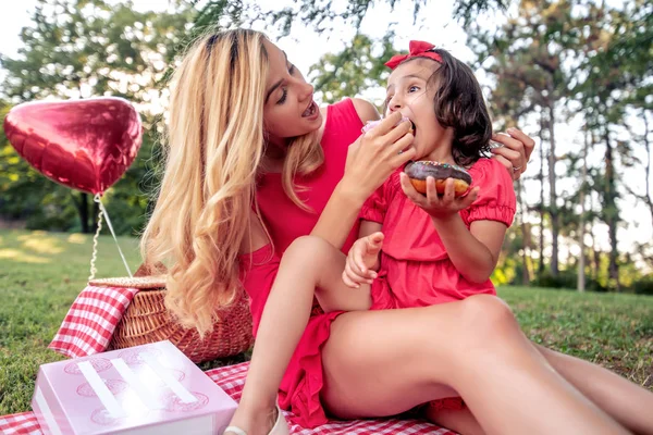 若い母親と彼女の娘は 公園で毛布の上に座っている間 ドーナツを食べて 楽しみを持っています — ストック写真
