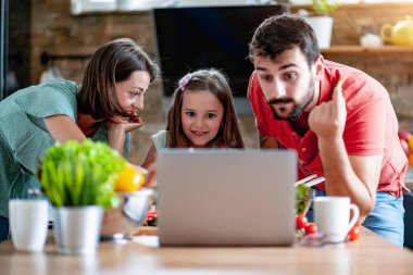 Yemek, yemek ve ev konsepti. Anne, baba ve çocuk mutfakta salata yapmak için sebze kesiyorlar. Eğleniyorlar ve tarifi internetten izliyorlar..