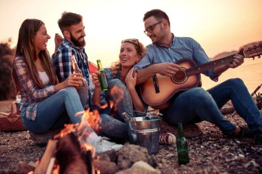 Bir grup heyecanlı genç arkadaş sahilde birlikte eğleniyor, bira içiyor, kamp yaparken gitar çalıyorlar.