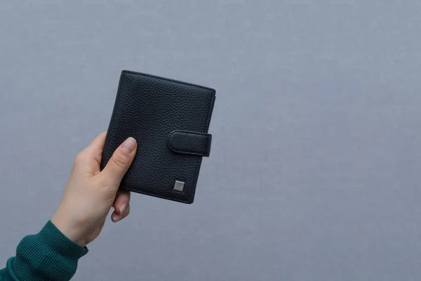 Young woman in green turtleneck holds large expensive wallet in her left hand. Wallet made of black genuine leather. Modern fashionable wallet on the gray background. Copyspace