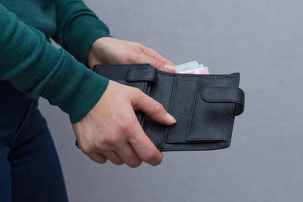 A woman in green turtleneck and blue jeans stands sideways on grey background. She takes some euro money out of good wallet of her husband. Modern wallet made of black genuine leather.