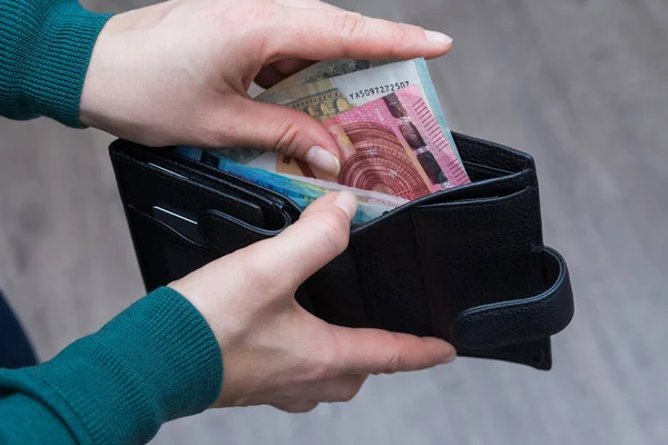 A woman in green turtleneck takes some euro money out of good wallet of her husband. Modern wallet made of black genuine leather.