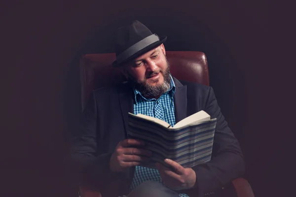 Portrait of man with beard and hat sitting on leather chair reading book. — Stock Photo, Image