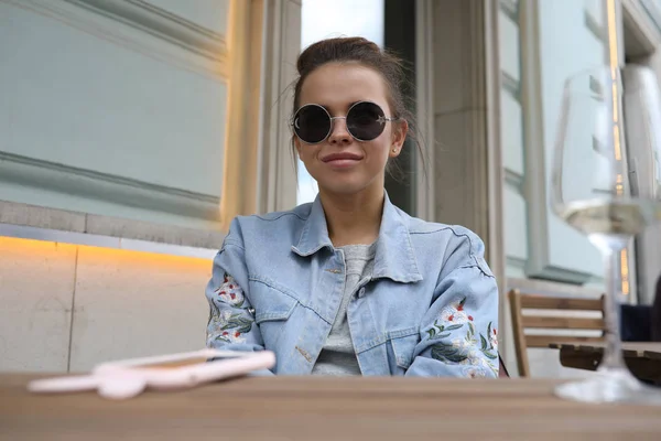 Cheerful girl in sunglasses sits at a table in a street cafe. — Stock Photo, Image