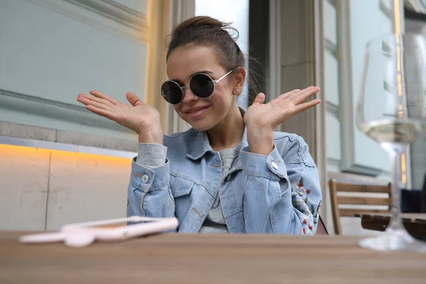 Cheerful girl in sunglasses sits at a table in a street cafe. — Stock Photo, Image