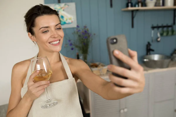 A woman drinks wine and makes selfie in the kitchen.