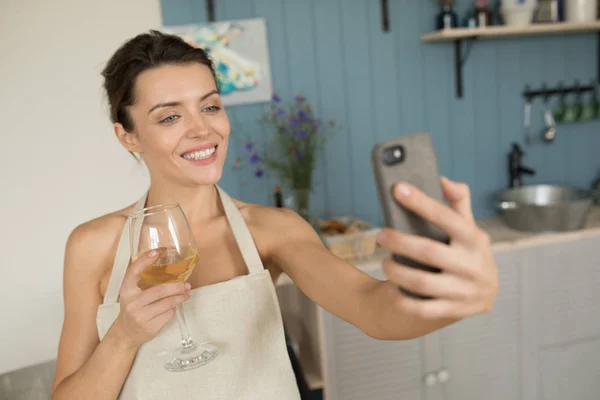 A woman drinks wine and makes selfie in the kitchen.